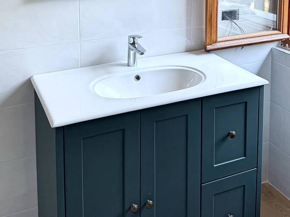 Bagno ristrutturato: lavabo bianco su mobile verde con ante a riquadri, accanto a una finestra in legno