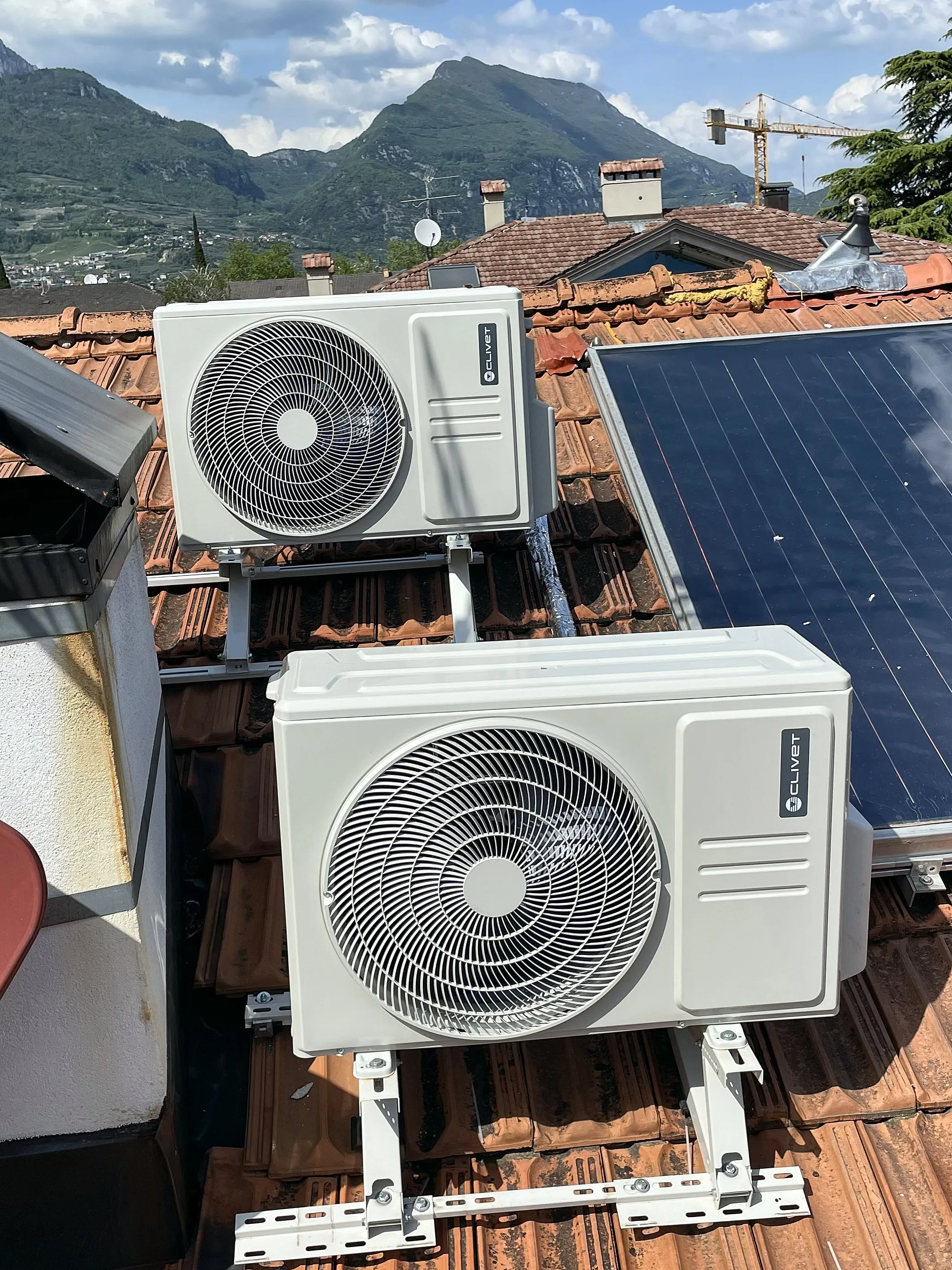 Due unità esterne di climatizzatori sulle staffe del tetto, accanto a un pannello solare, con le montagne dell'Alto Garda dietro