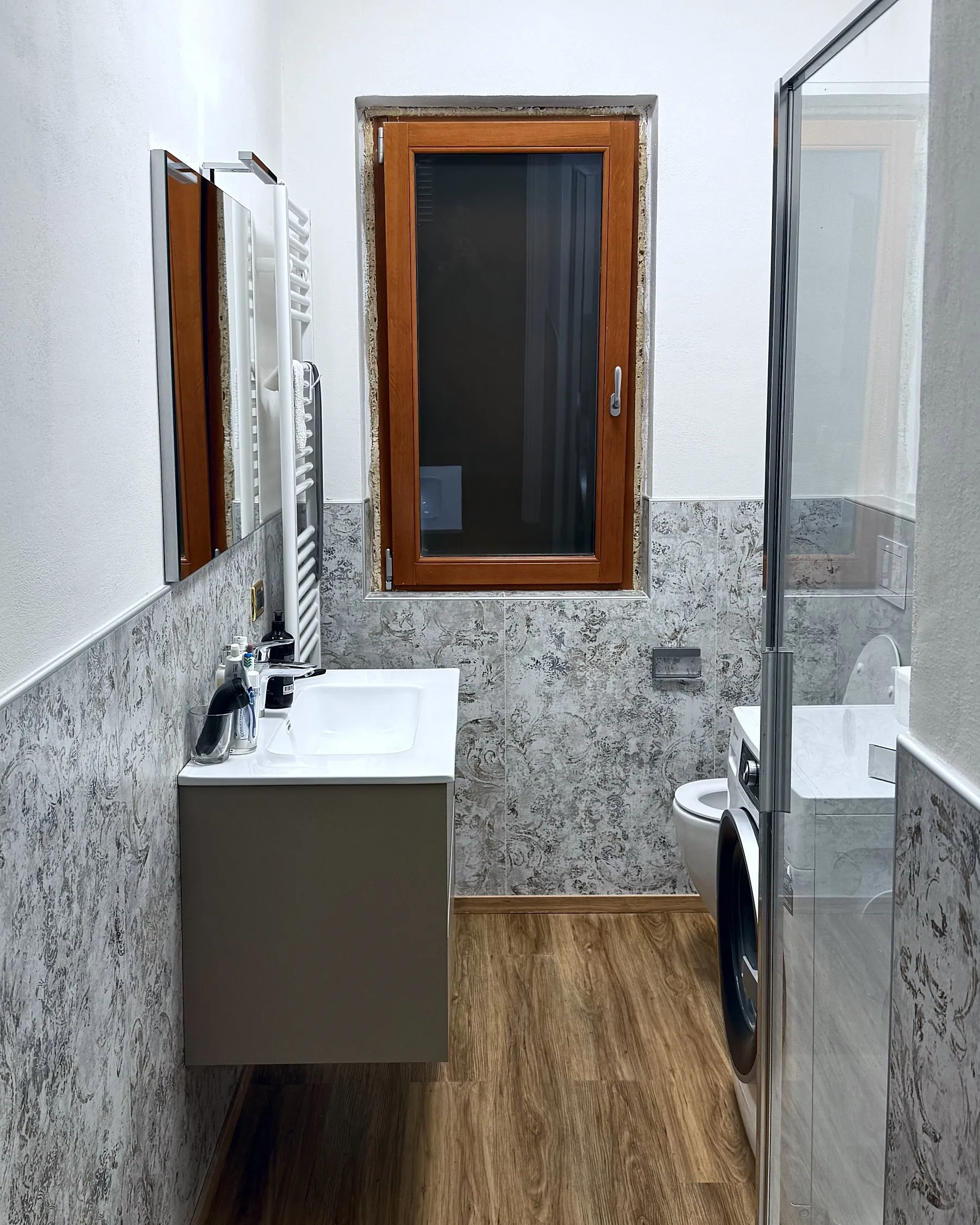 Bagno moderno e funzionale, con mobile lavabo sospeso, termoarredo, sanitari compatti e box doccia in vetro. Spazi ottimizzati per integrare anche la lavatrice, senza rinunciare al comfort e all'eleganza.