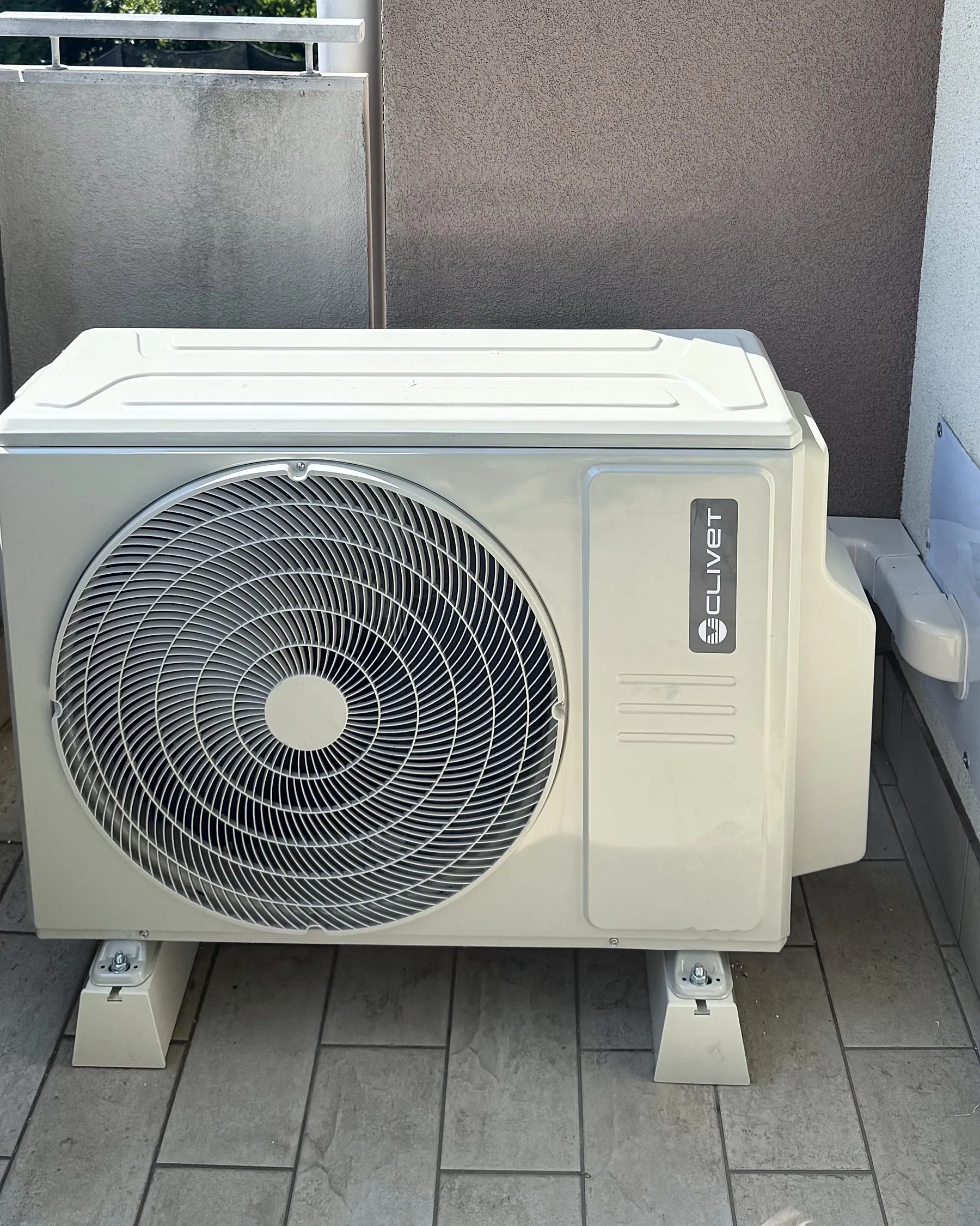 Unità esterna di un climatizzatore sul balcone, sui suoi piedini, con le montagne dell'Alto Garda oltre la ringhiera