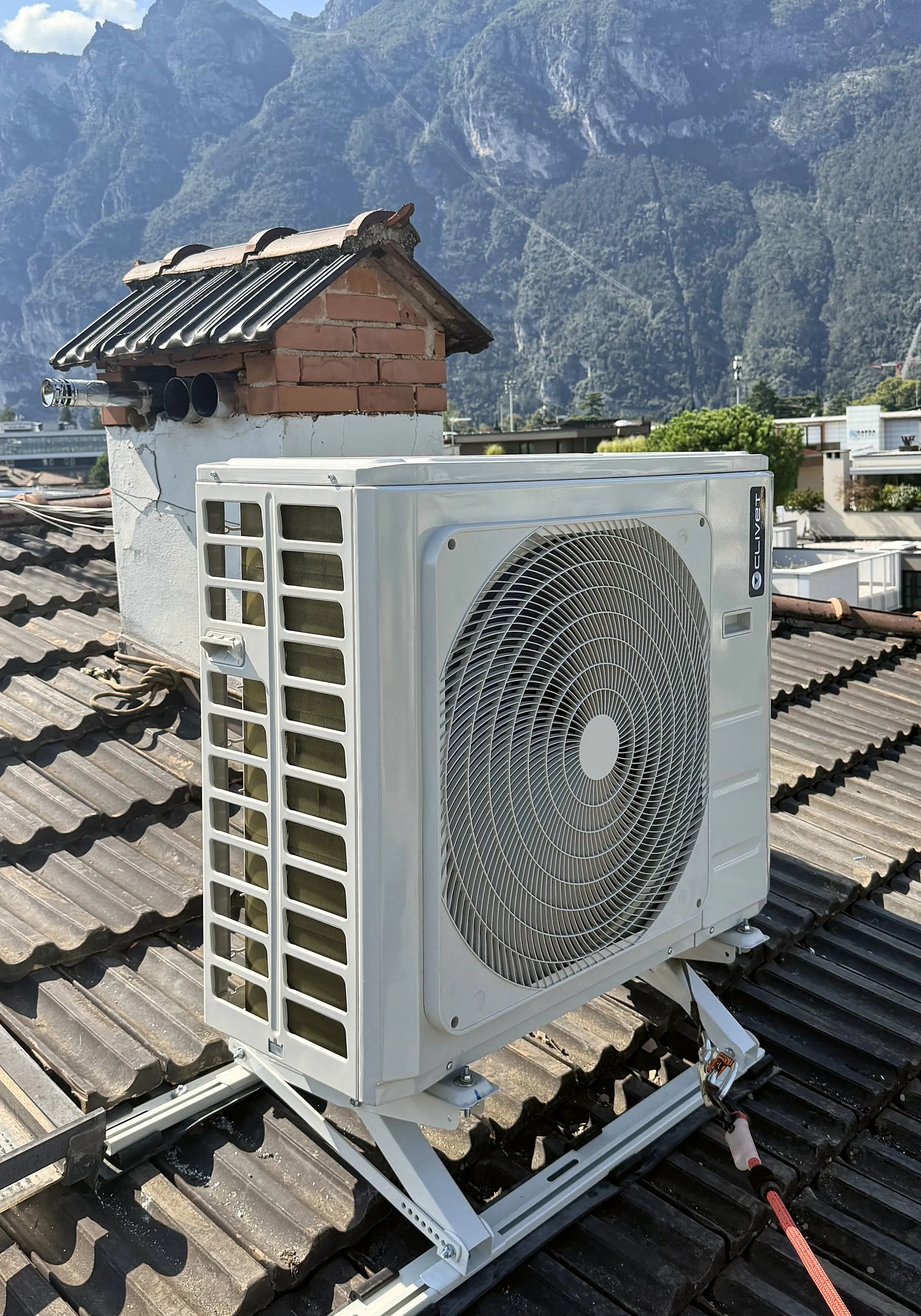 Unità esterna di un climatizzatore sulle staffe di un tetto in coppi, con le montagne dell'Alto Garda e il cielo dietro